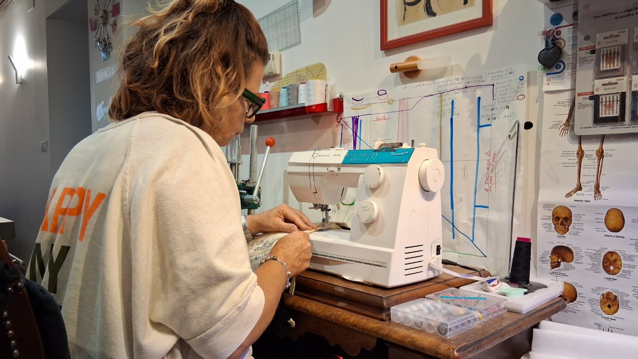 La praticienne à la machine à coudre de l'atelier, devant un patron de ceinture affiché au mur et une planche d'anatomie.