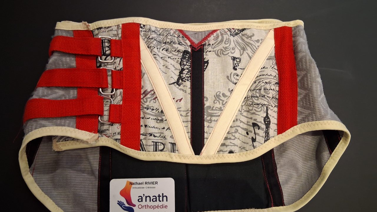 Une ceinture lombaire sur mesure terminée, en tissu imprimé gris et blanc, avec ses sangles de rappel rouges et une carte de visite a'nath-Orthopédie.