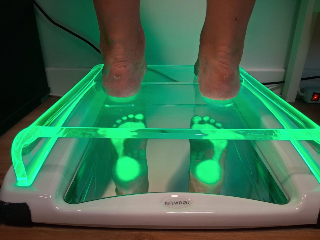Deux pieds nus posés sur un podoscope éclairé en vert : le miroir montre les zones de la plante qui portent le poids.