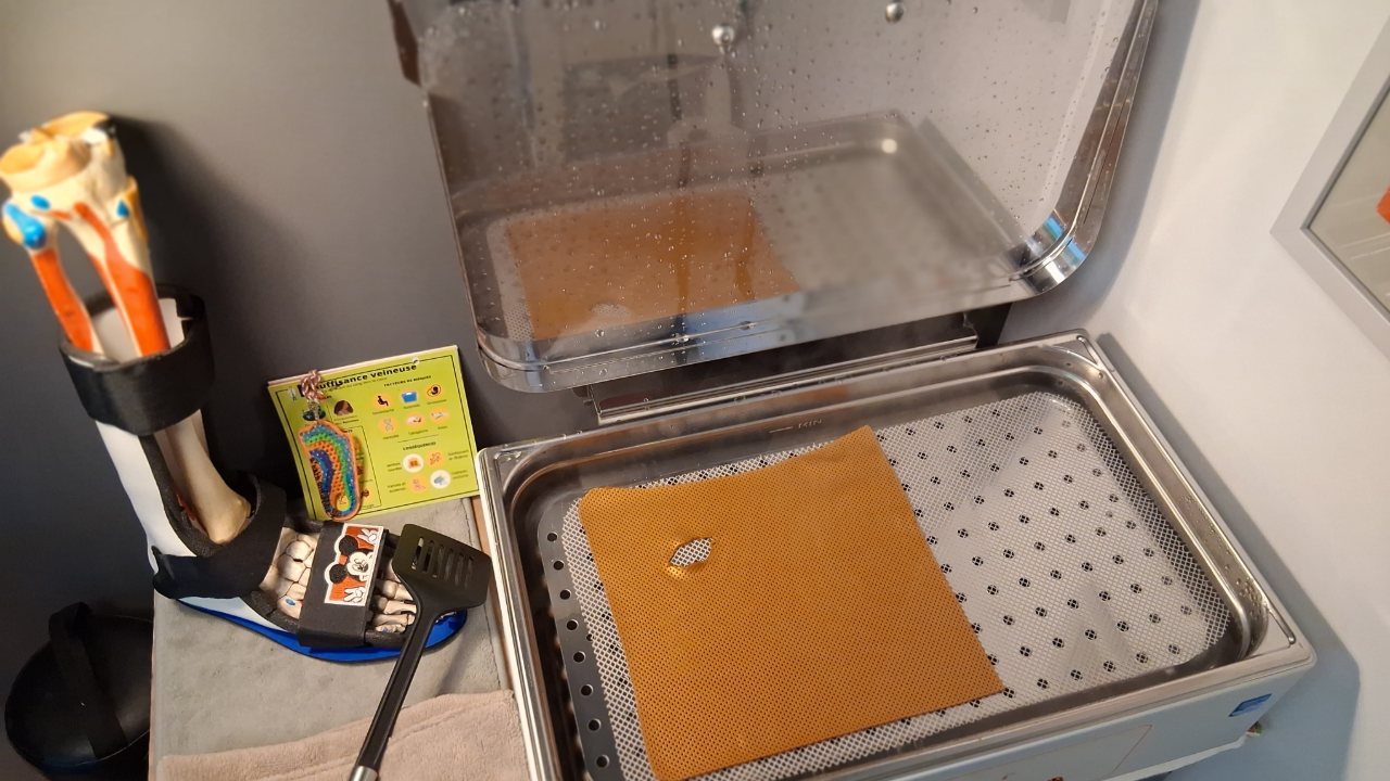 Le bac d'eau chaude du cabinet, ouvert, avec une plaque de thermoplastique orange perforée en train de ramollir ; à gauche, un releveur de pied sur un modèle anatomique.